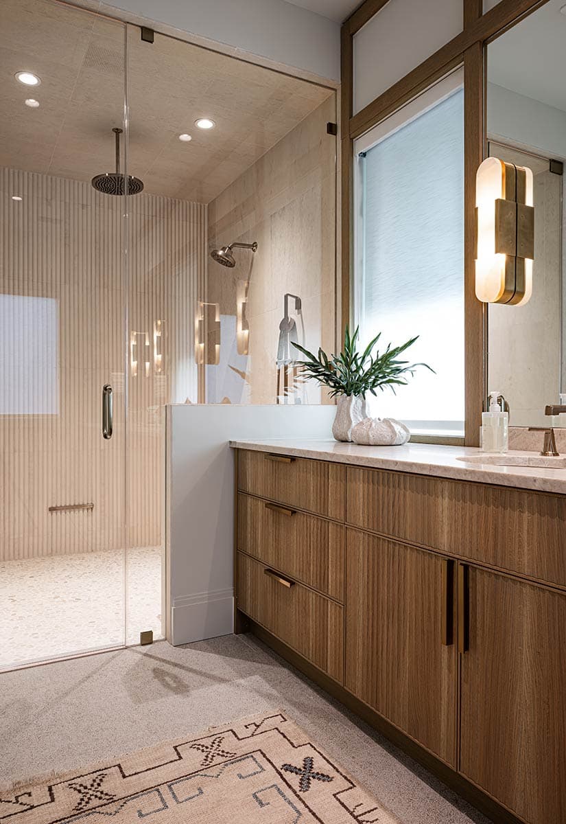 A modern bathroom showcases a glass shower, wooden vanity, marble countertop, and a decorative rug on the floor. A plant sits on the vanity.