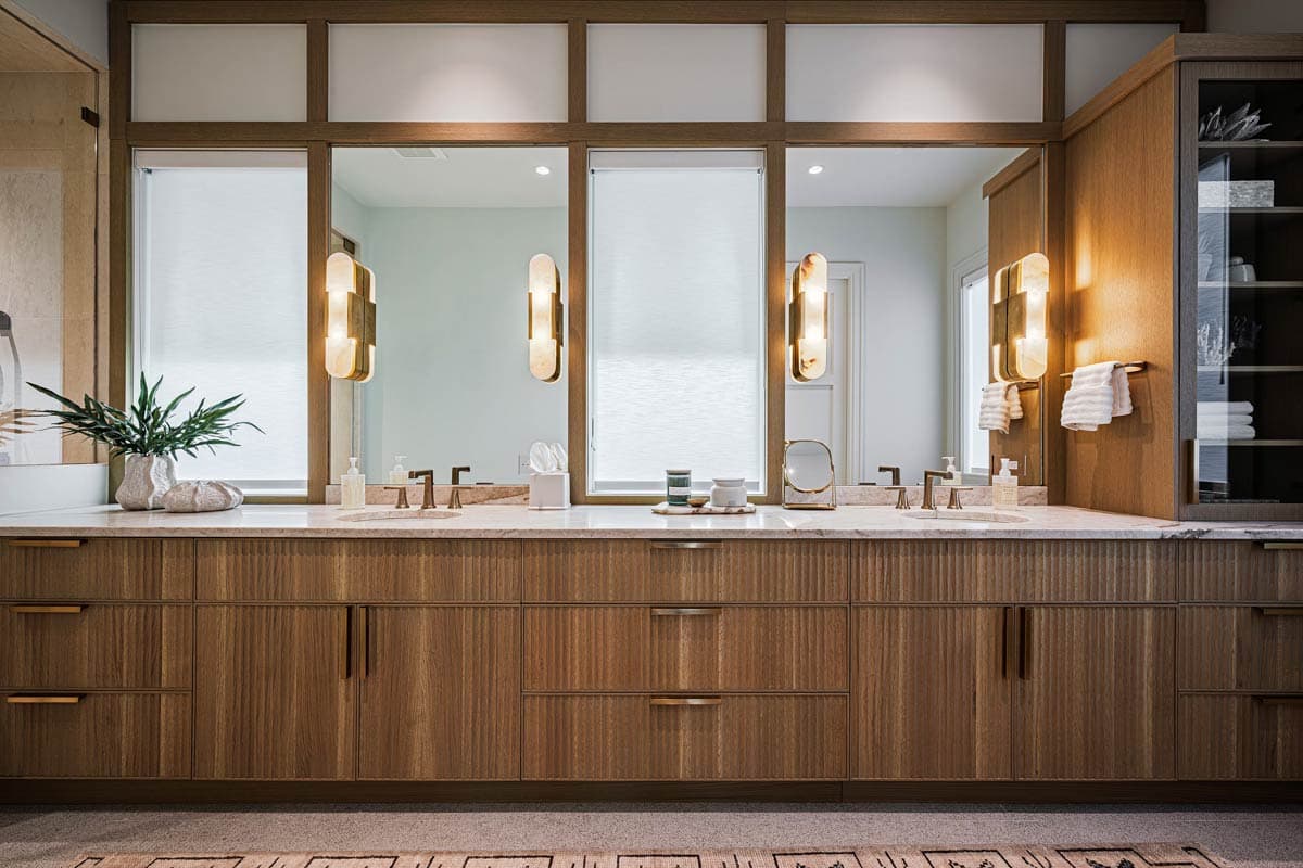 A modern bathroom vanity with a wood grain cabinet, marble countertop, and illuminated sconces flanking mirrors. Includes a linen cabinet.