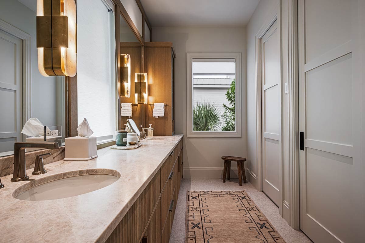 A modern bathroom showcases a marble countertop with sinks. Wooden cabinets and a decorative rug complete the room. A window offers an outdoor view.