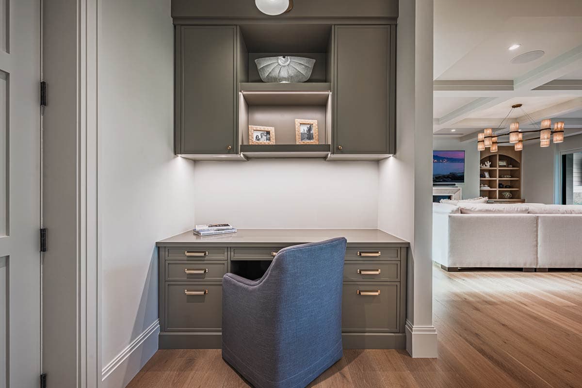 A built-in desk with grey cabinets and a blue chair sits in a hallway, offering a view of a living room in the background.