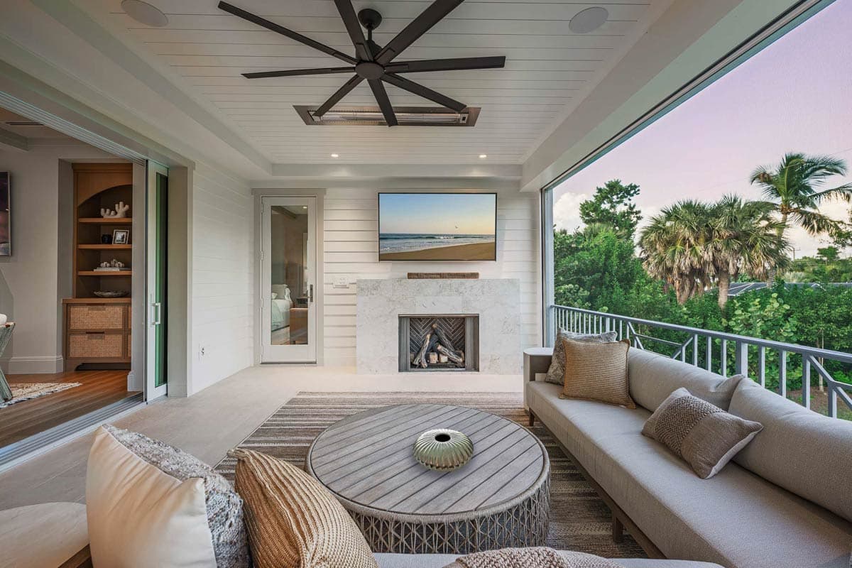 A modern outdoor living space features a fireplace, TV, and large ceiling fan. A large open window reveals a tropical view and a gray sofa.