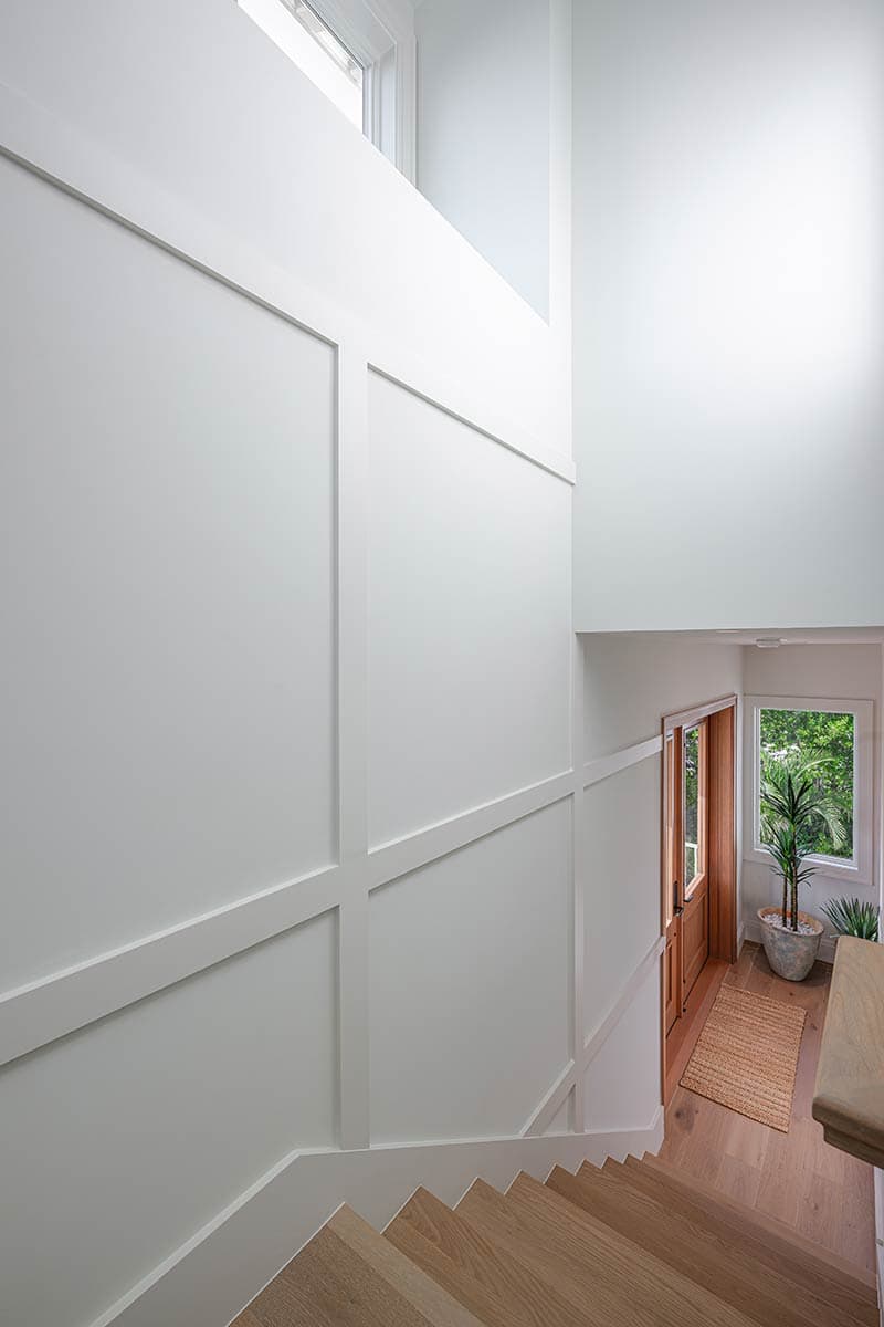 Looking down a stairwell with white paneled walls. A wooden door and window open to a bright, green view at the bottom of the stairs.