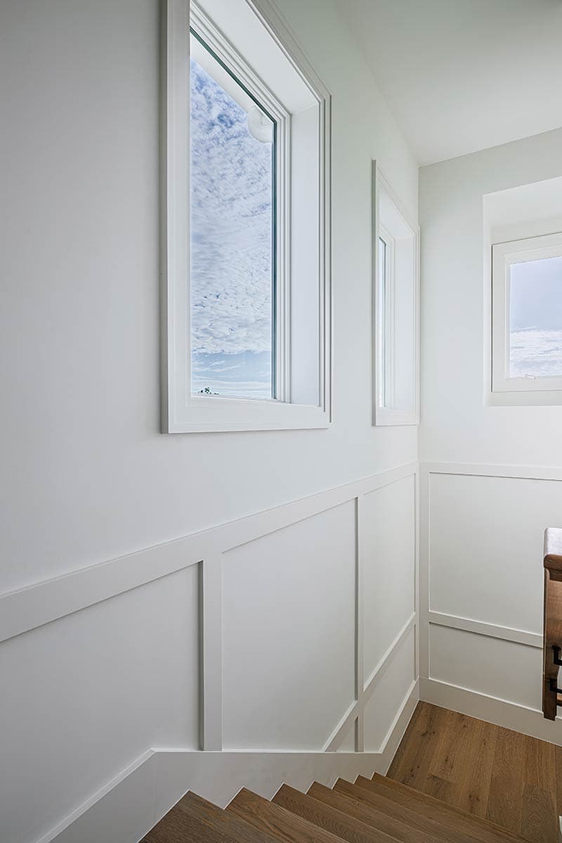 Bright hallway with white walls and trim, punctuated by windows framing a sky view. Wooden stairs lead downwards on the right.