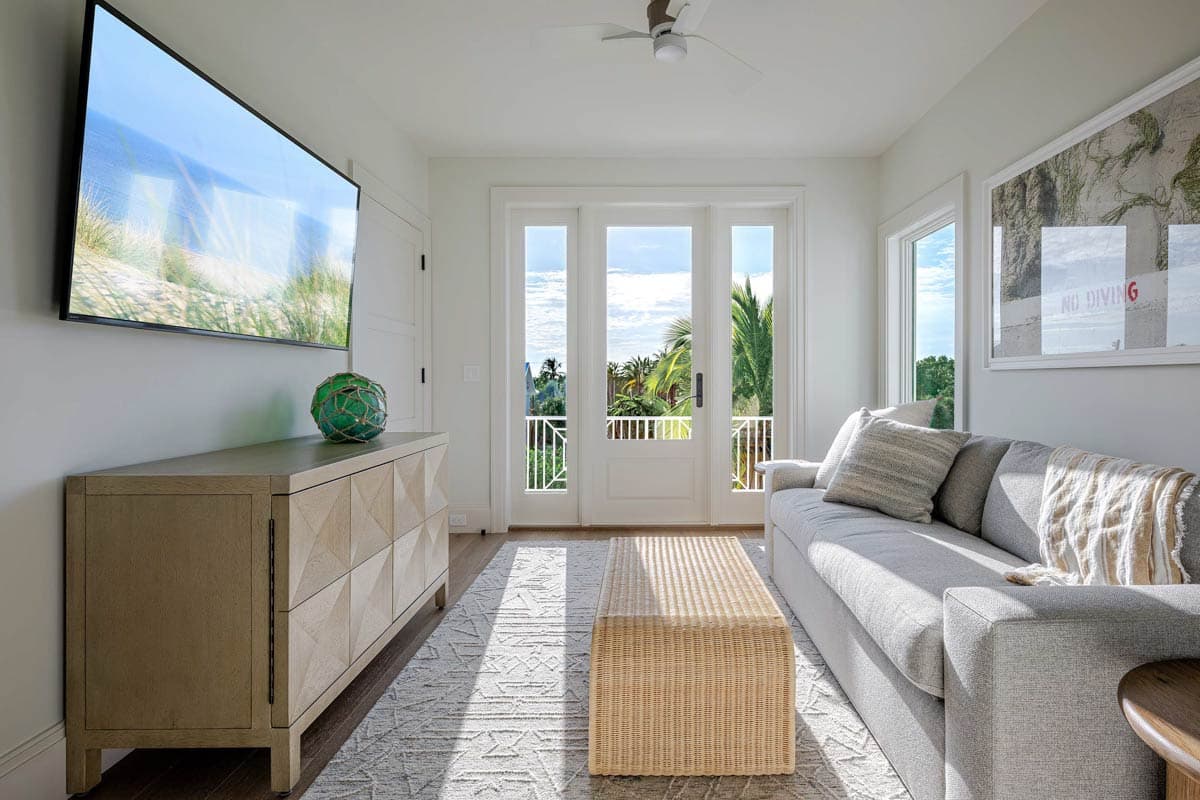Bright, modern living room with a sofa, woven coffee table, and large TV. Doors open to a balcony with palm trees visible outside.