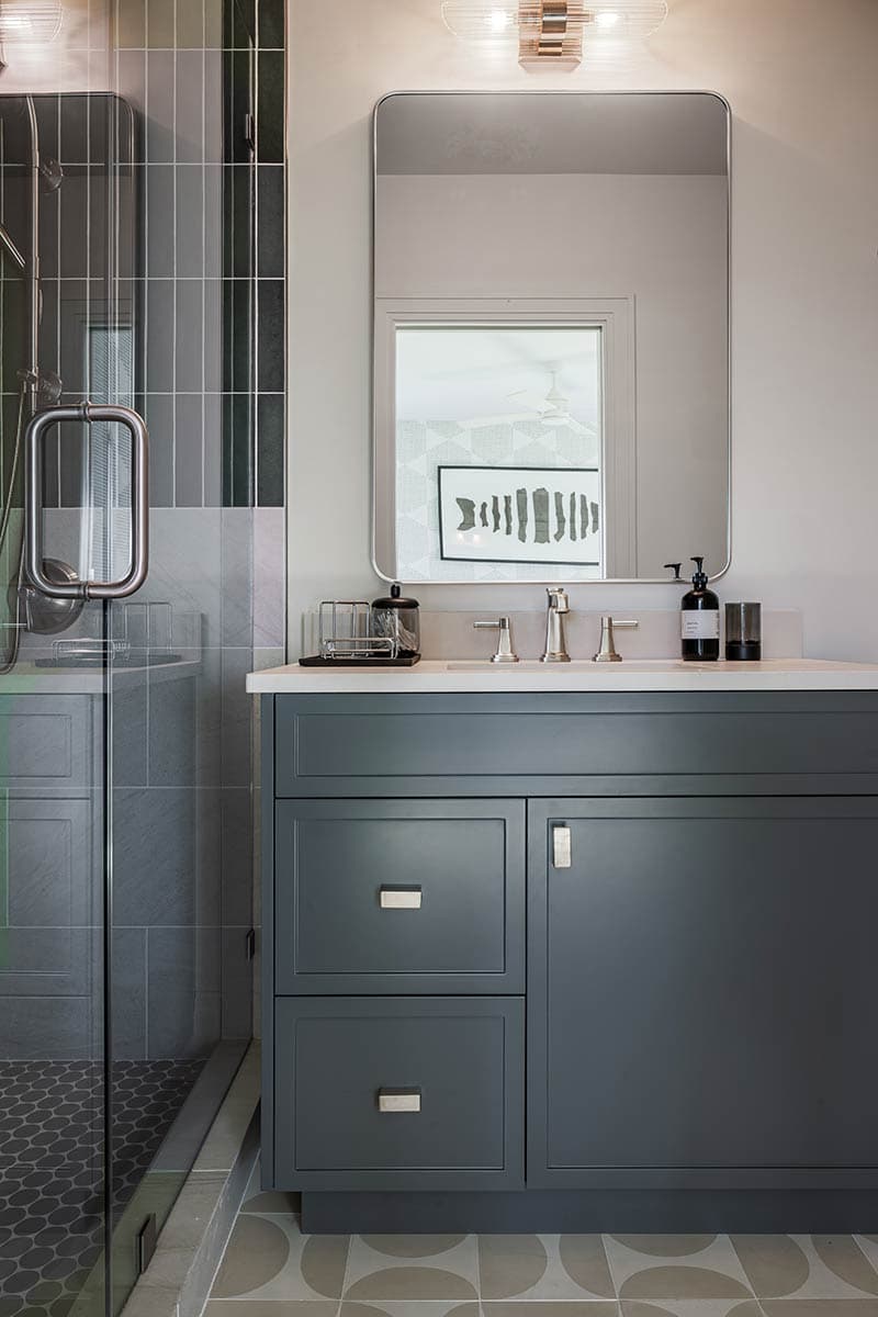 A modern bathroom vanity features a dark gray cabinet, rectangular mirror, and a glass-enclosed shower on the left side.