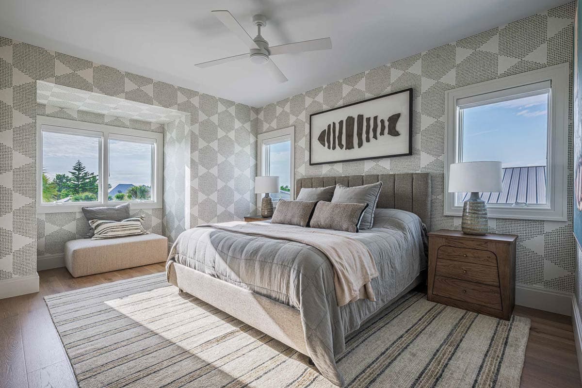 Bright bedroom with patterned wallpaper, large bed, and window seat. A fish art piece hangs above the bed, with striped rug below.