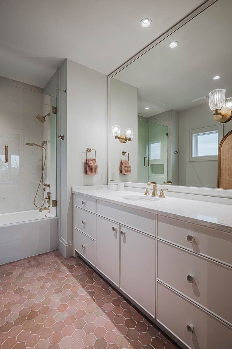 A modern bathroom with white cabinets, a large mirror, and rose-colored hexagon tile flooring. Gold fixtures and a glass shower complete the space.