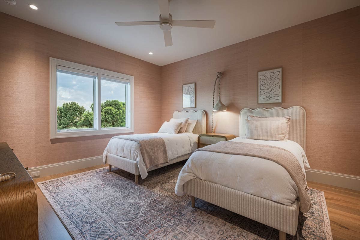 A bedroom features two twin beds, decorative wall art, and a large window overlooking trees. Pink textured wallpaper and area rug complete the look.