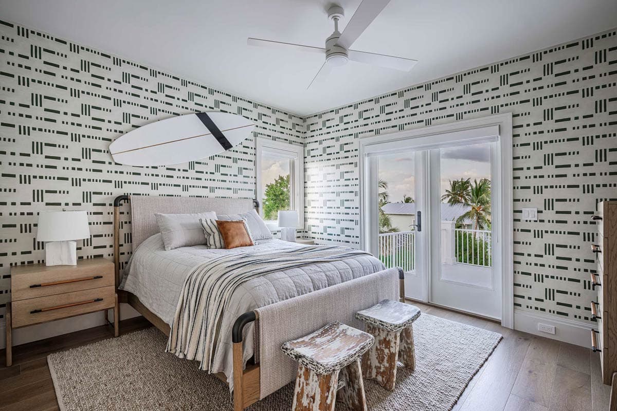 A modern bedroom with patterned wallpaper, a bed, and a surfboard mounted on the wall. A balcony is visible through glass doors.