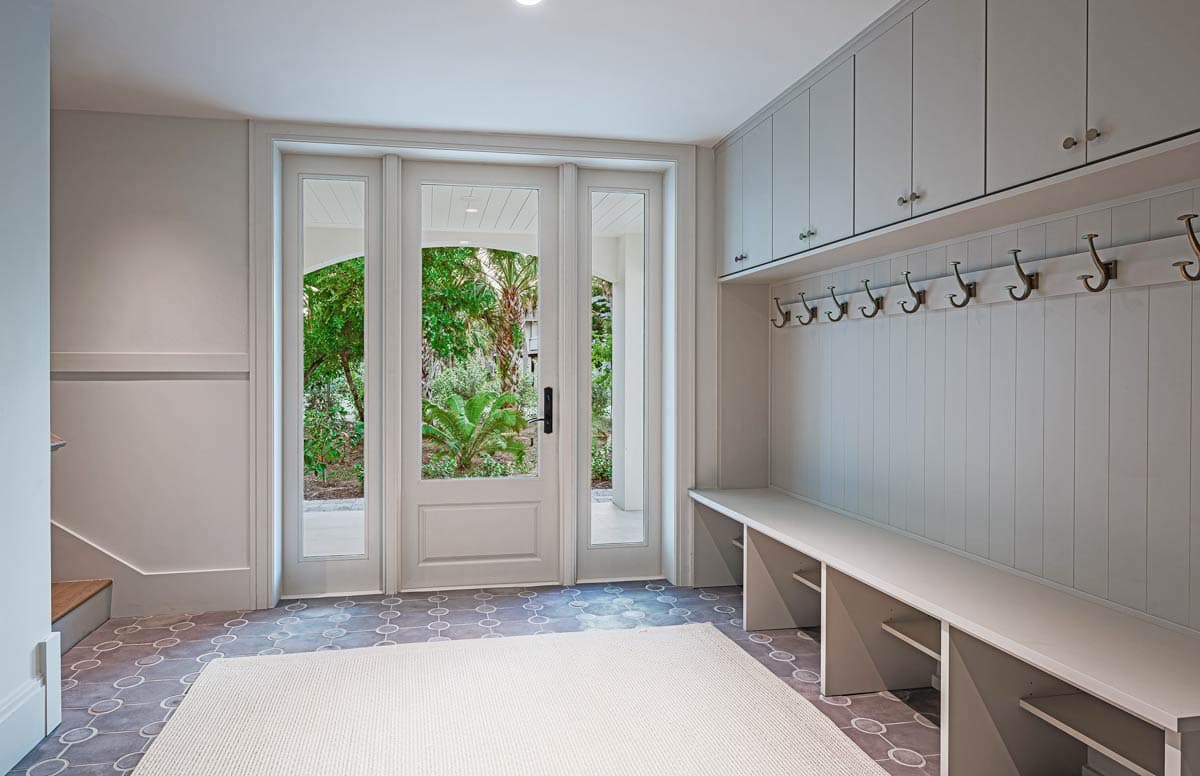 Bright entryway with white paneled walls, bench with hooks, and a glass door leading to a lush green outdoor area.