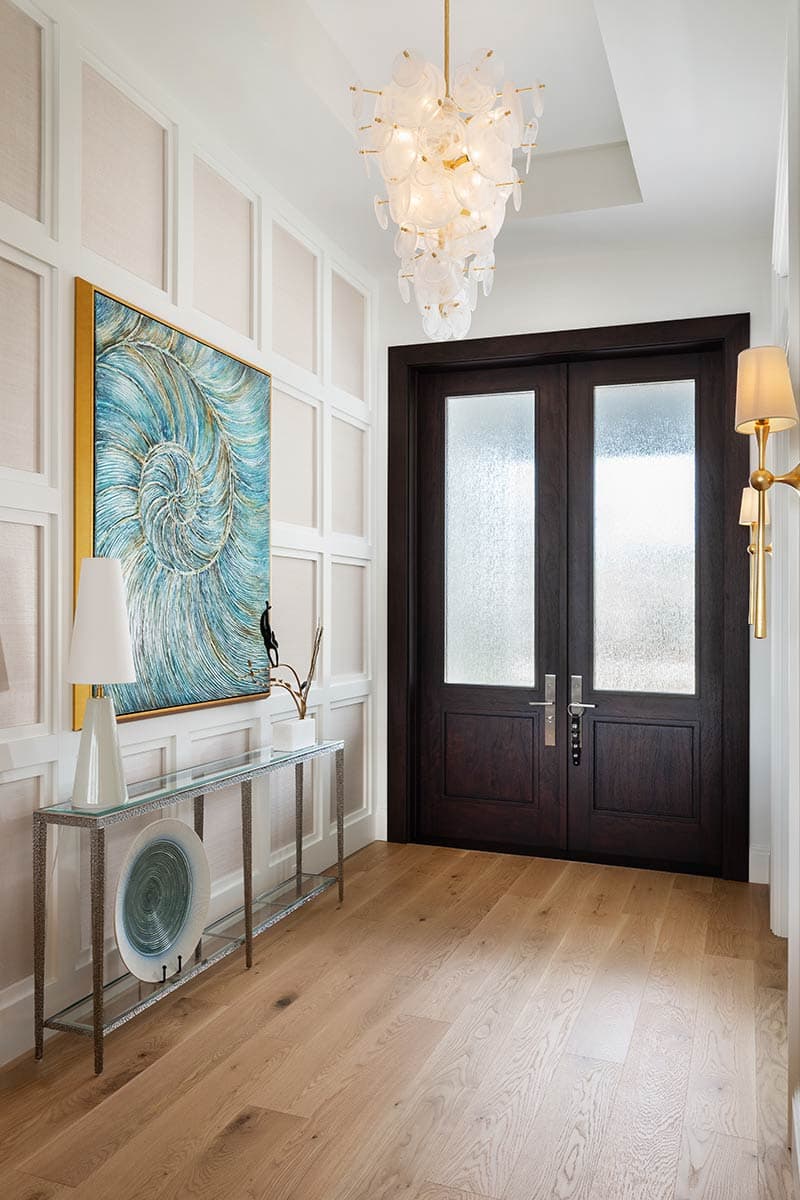 Interior view of a hallway with double dark wood doors, textured glass panes, wainscoting, and a glass console table.