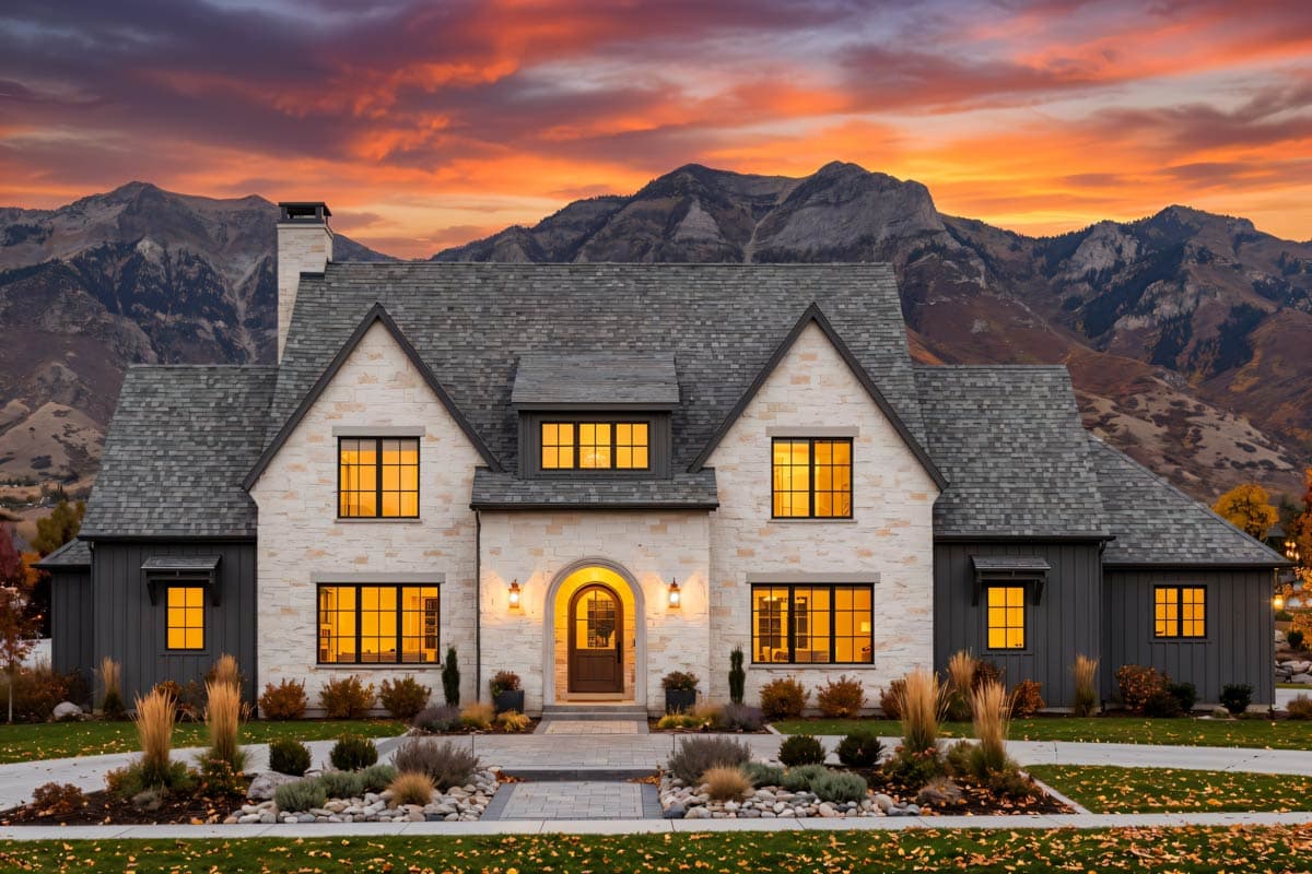 A large, stone-clad house glows with warm interior lights as dusk settles, with mountains in the background under a vibrant sunset sky.