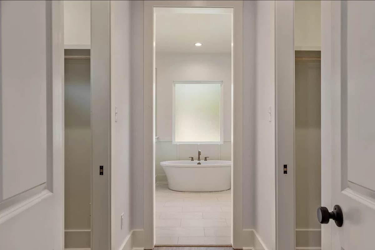 View through doorways into a bathroom with a freestanding tub and frosted window. Open closets flank the hallway.