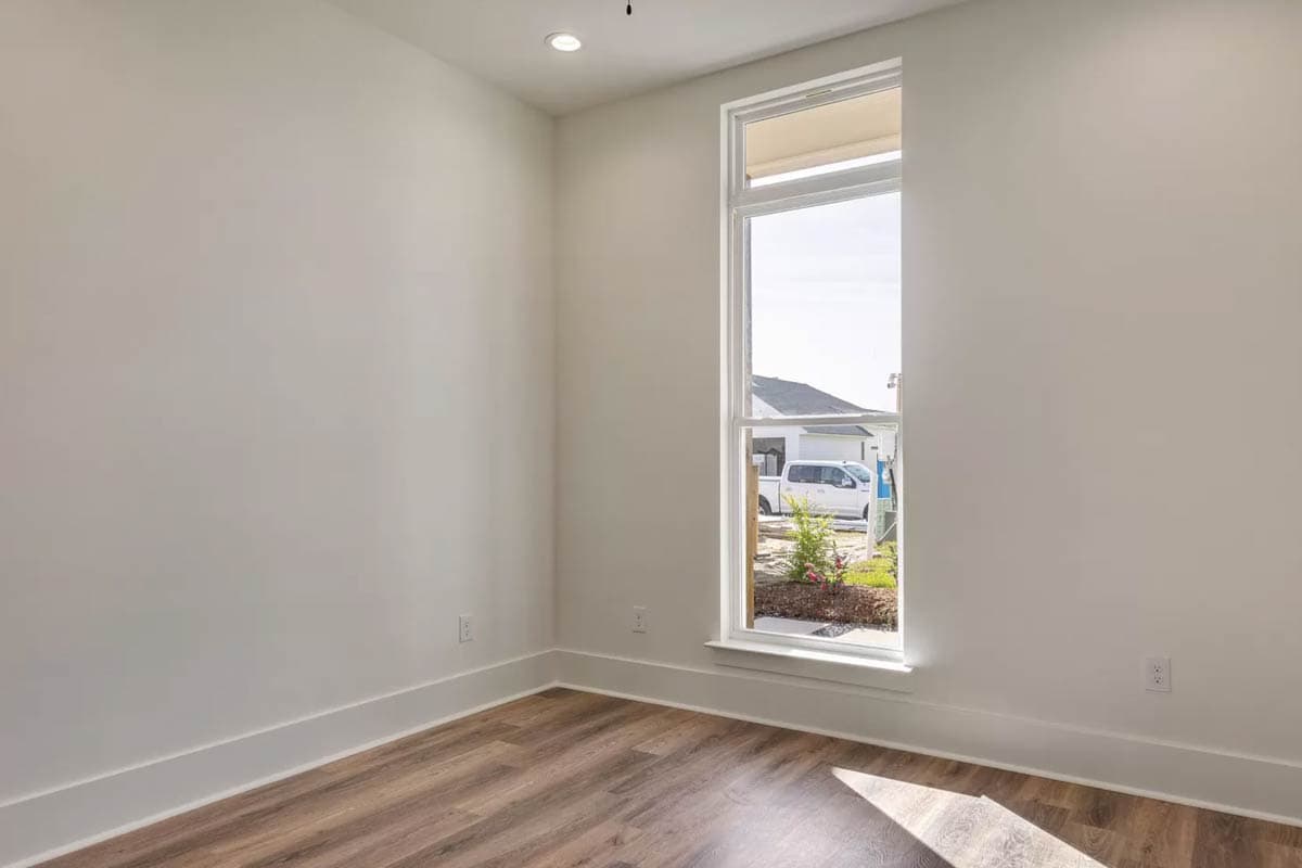 Interior view of an empty room with wood-look flooring and a tall, narrow window overlooking a street with houses and a white truck.