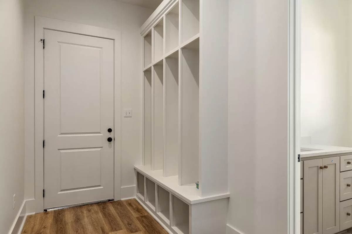 Interior view of a mudroom with a built-in white bench and cubbies next to a paneled door. A vanity is visible through an opening.
