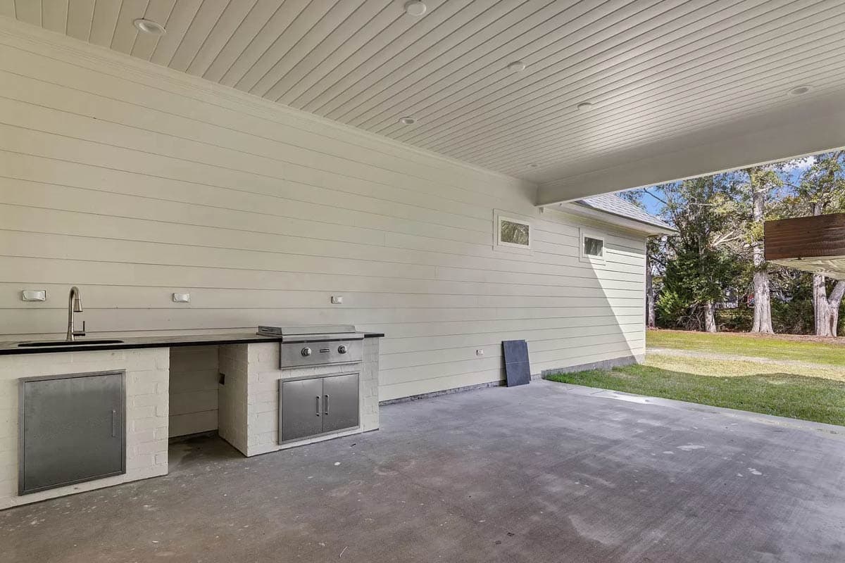 Outdoor kitchen with sink, built-in grill, and storage cabinets on concrete patio under a covered ceiling.