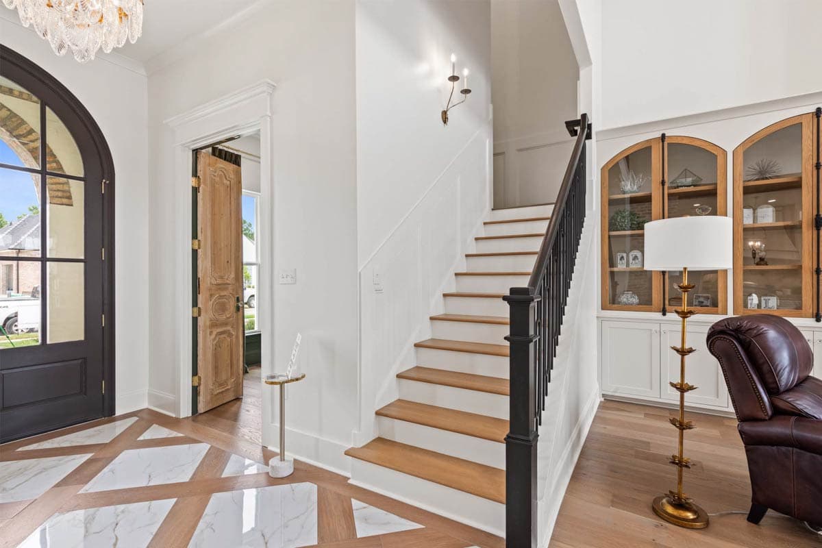 Interior view of a foyer with a grand staircase, arched doorway, marble and wood inlaid floor, and built-in display cabinets.