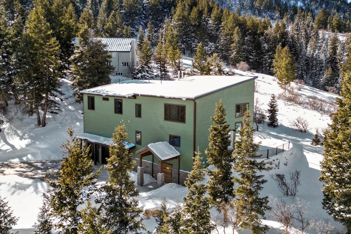 Modern house plan exterior with green siding, multiple gables, covered entry, and large windows, set in a snowy, wooded landscape.