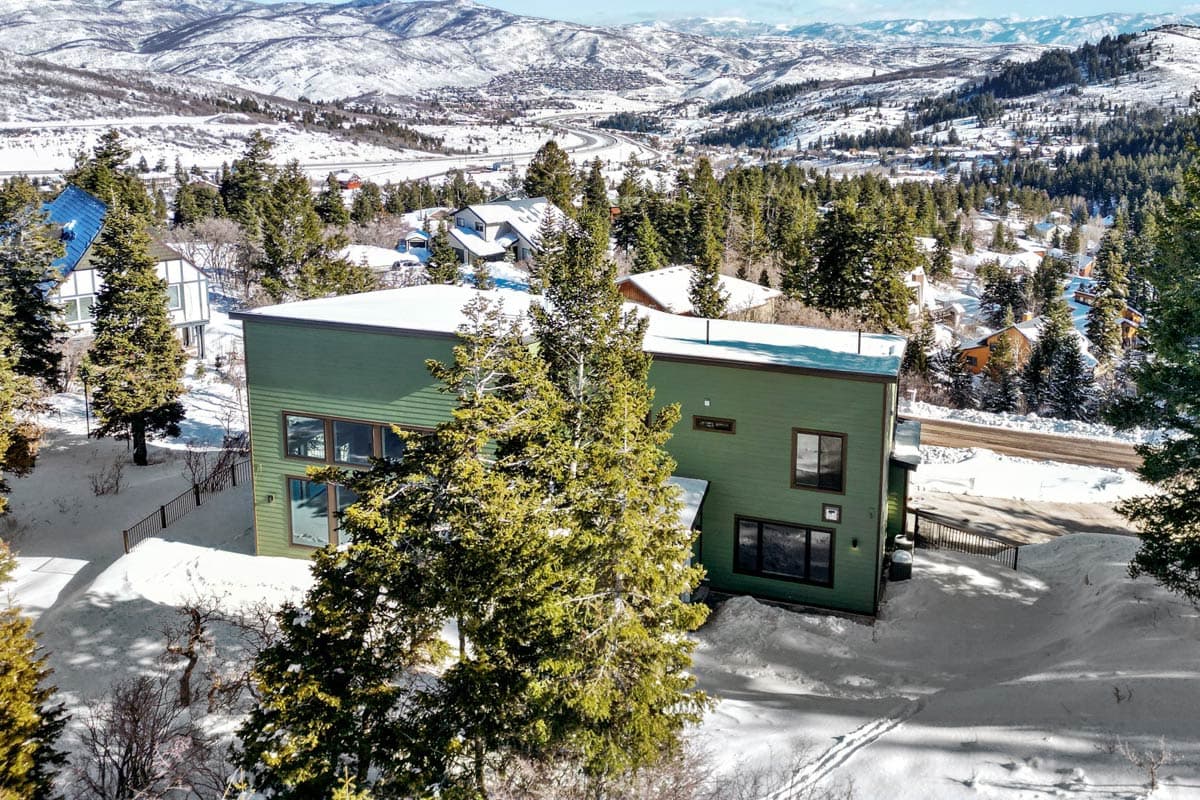 Modern house plan exterior with green siding, a flat roof, and large windows, set in a snowy, mountainous landscape.