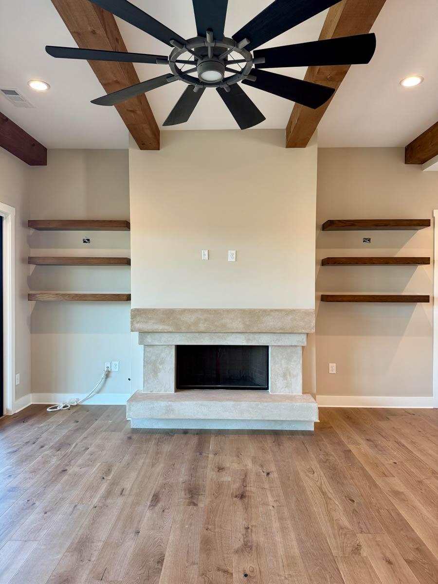 Living room with stone fireplace, floating shelves, wood beams on ceiling, and large ceiling fan.