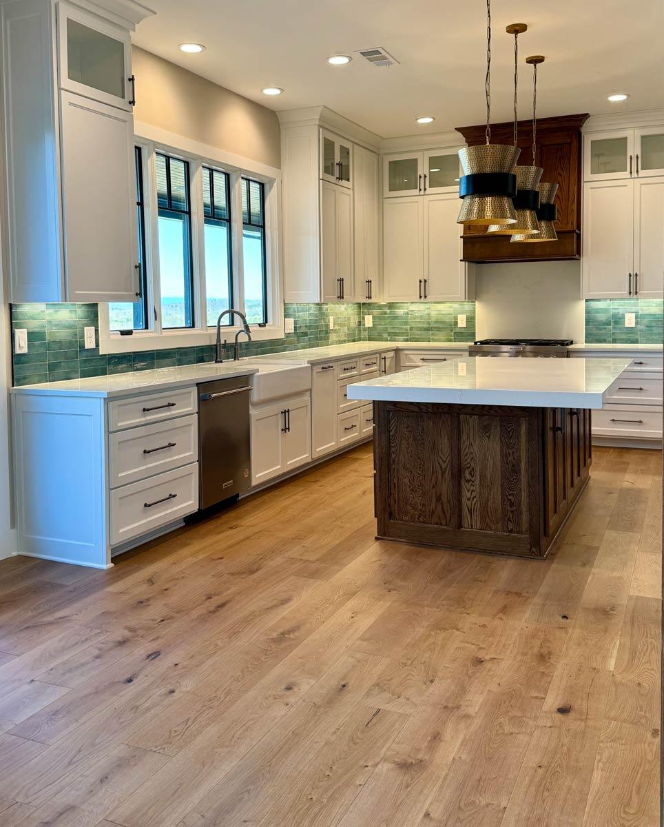 Kitchen interior with white cabinetry, large island, green tile backsplash, farmhouse sink, and pendant lighting.