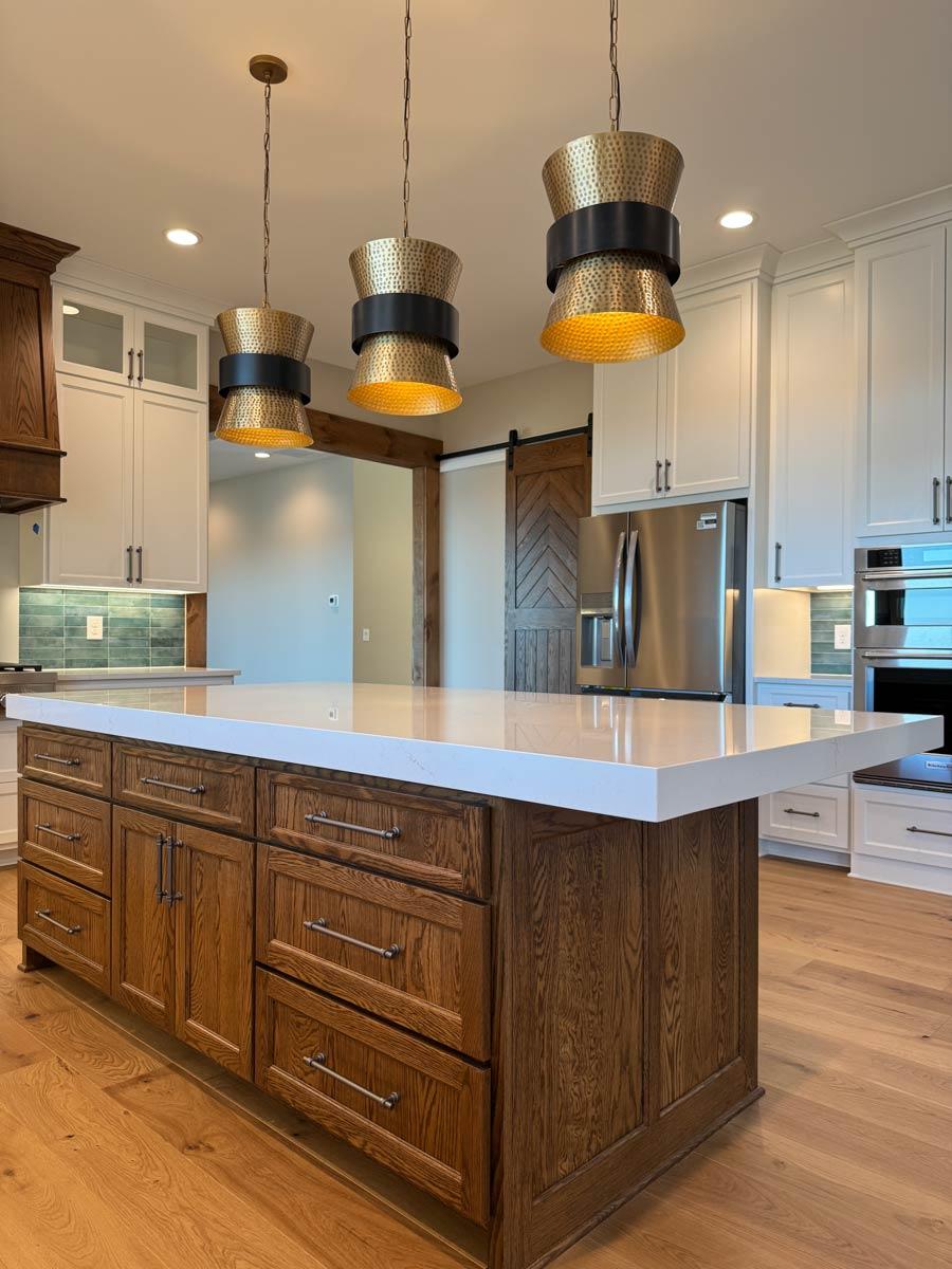 Kitchen island with white quartz countertop and stained wood cabinetry, white upper cabinets, stainless steel appliances, and pendant lights.