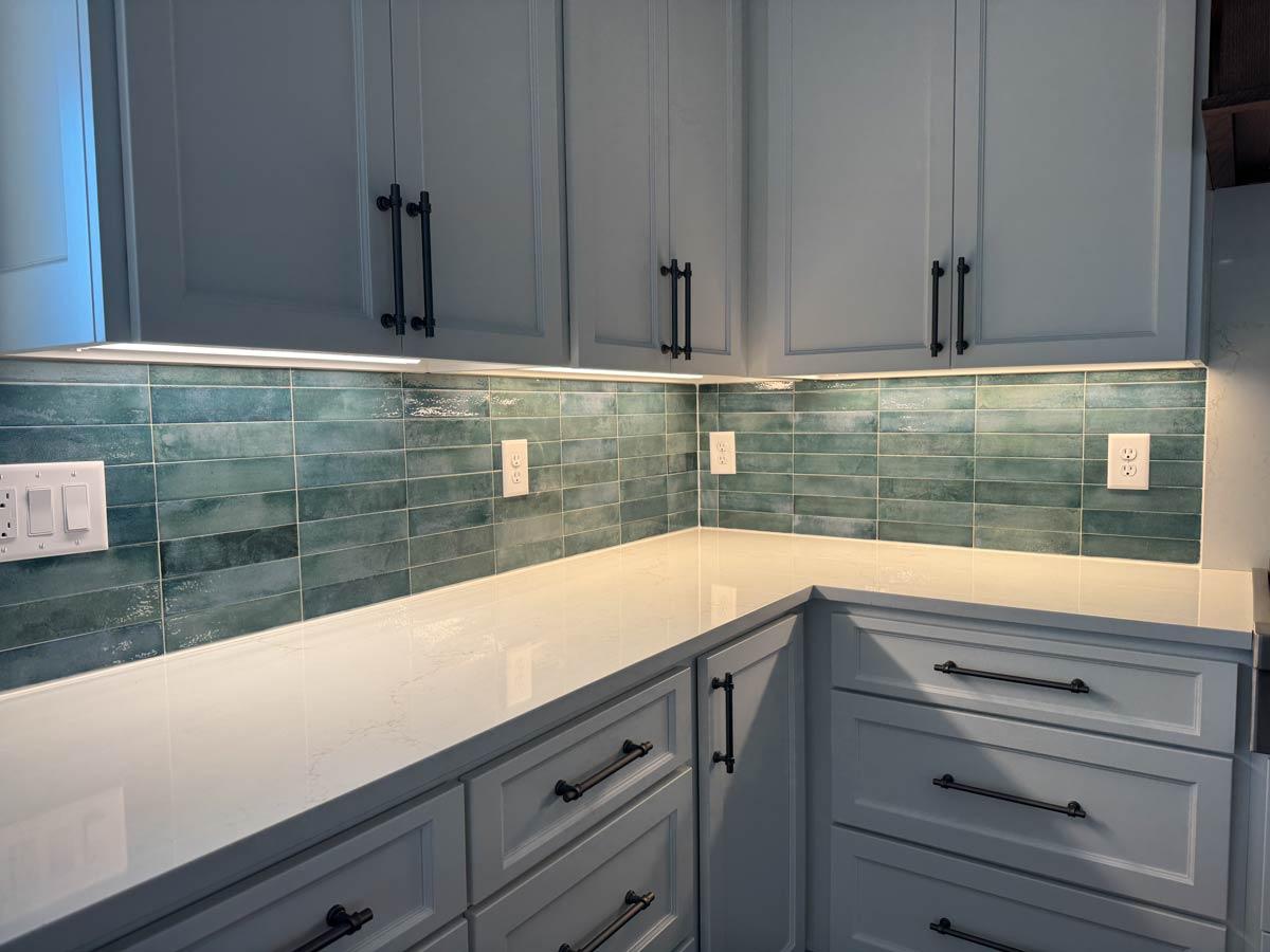 Kitchen corner with light gray cabinets, white quartz countertops, and teal subway tile backsplash with under-cabinet lighting.
