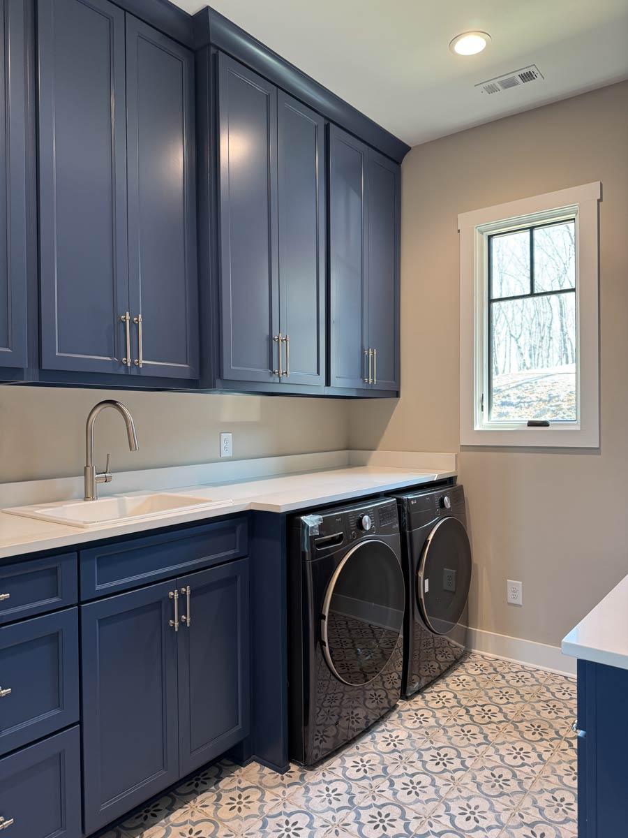 Laundry room with navy blue cabinets, white countertops, white sink, and two black washers/dryers. Patterned tile floor.