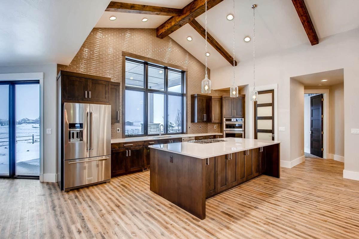 Modern kitchen with a large island, vaulted ceiling with exposed beams, and expansive windows. Stainless steel appliances and dark wood cabinetry.