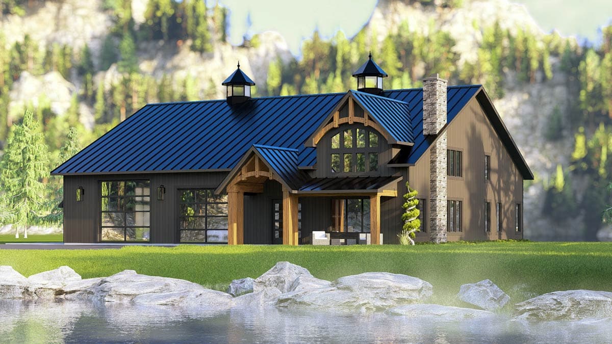 A modern cabin-style house with a dark blue roof and stone chimney sits in front of a mountain and a lake with large rocks.