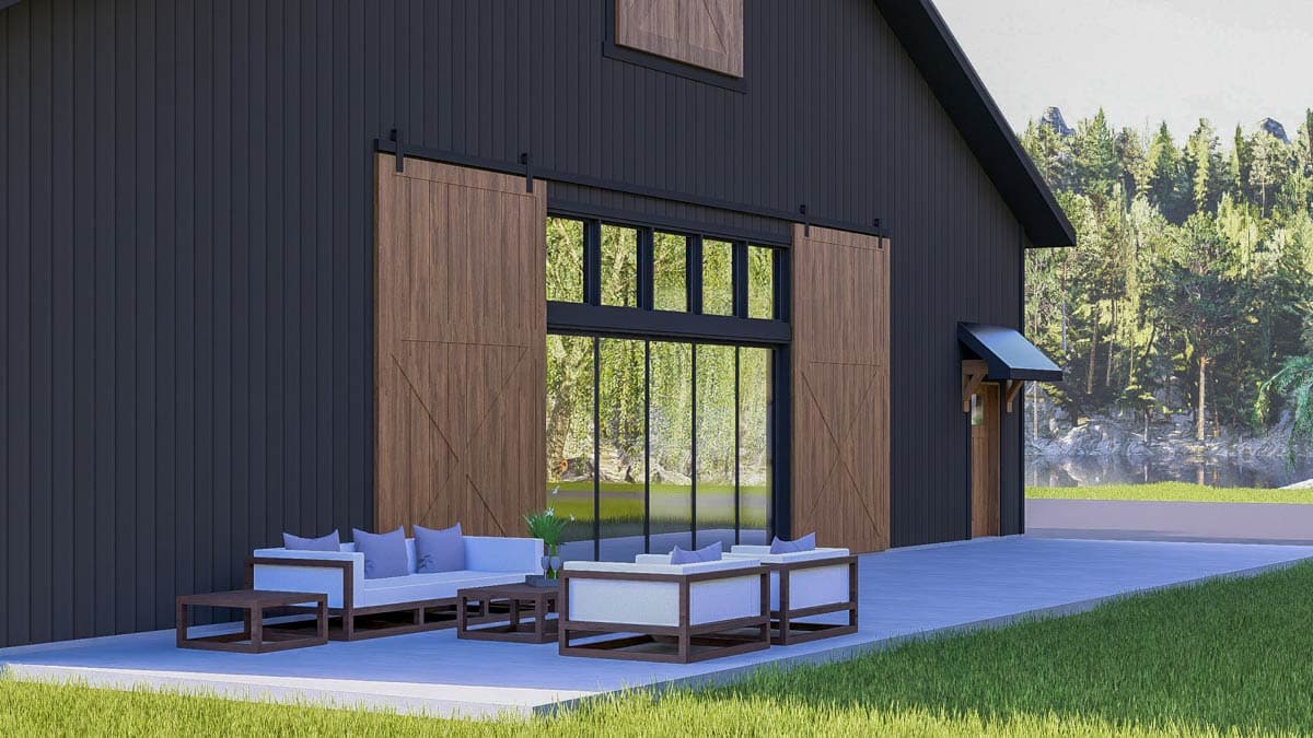 Modern black barn house with large wooden sliding doors, outdoor seating, and a large window overlooking a lush landscape and lake.