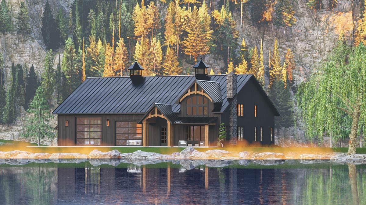 A modern cabin with a dark exterior sits beside a serene lake, reflecting the structure and surrounding forest of trees on a mountainside.