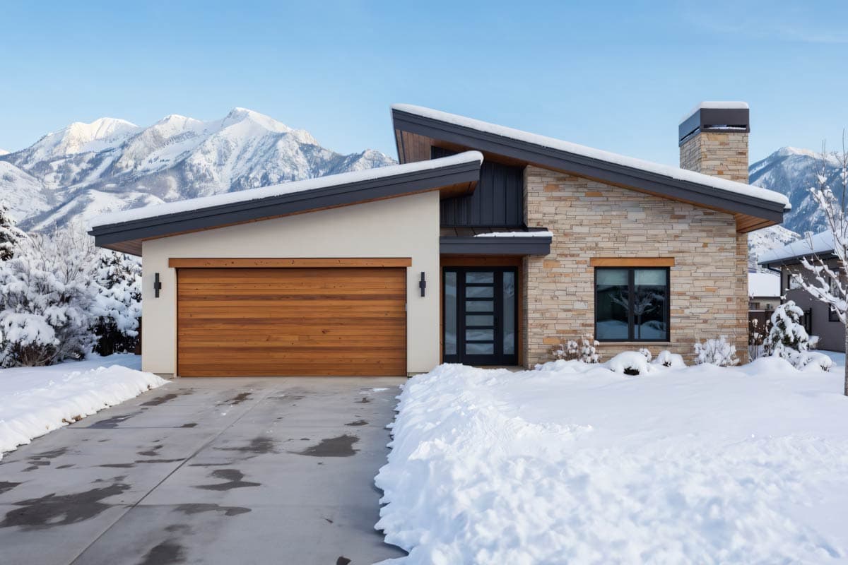 Modern house plan exterior with pitched rooflines, stone facade, wood garage door, and glass entry door, set against a snowy landscape.