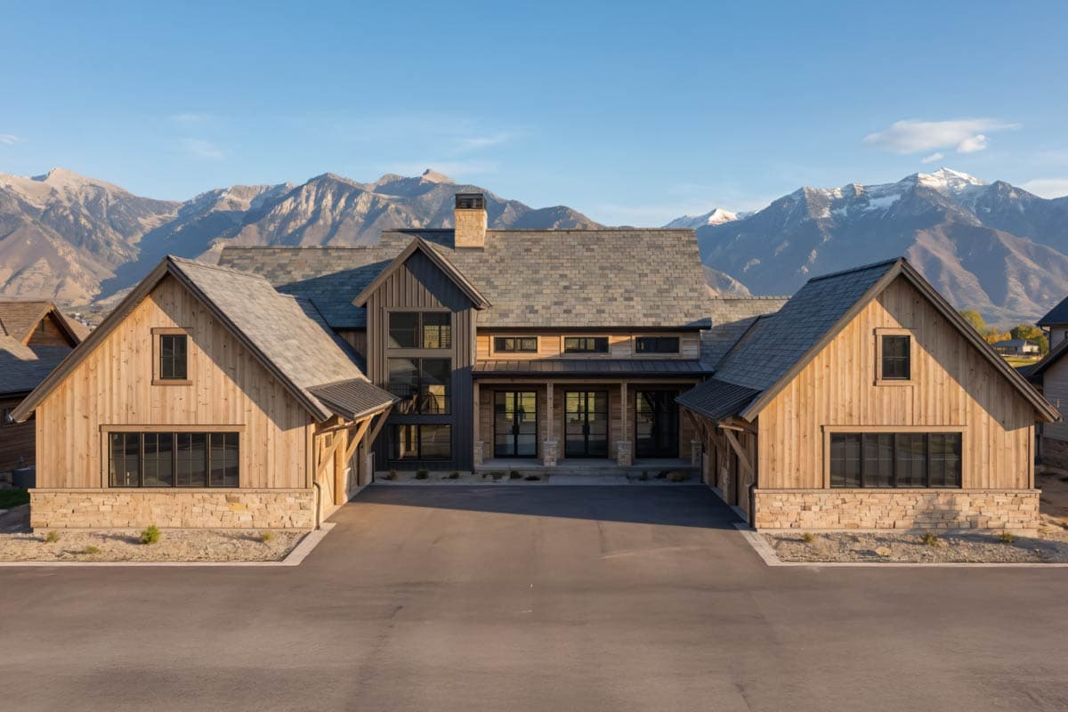 Sprawling modern home with wood siding and stone accents, flanked by two garage wings, with mountain range in the background.