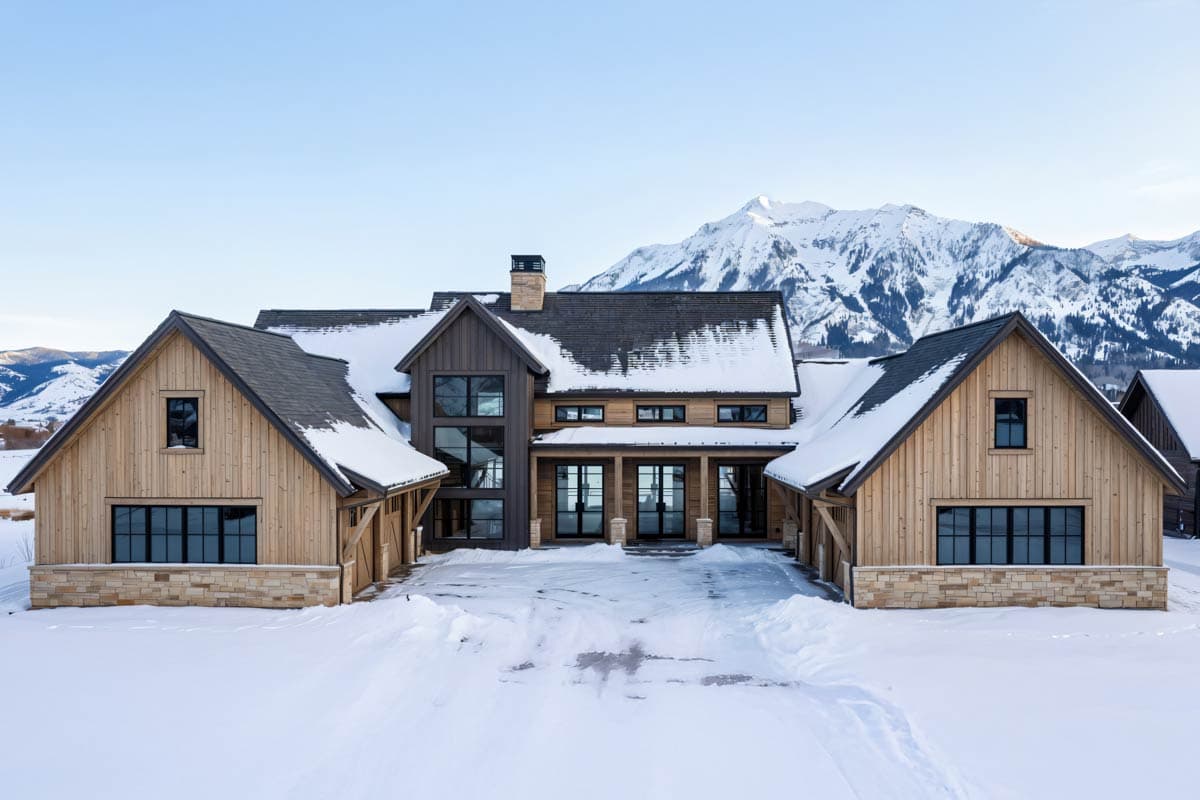 A modern, wood-sided home sits in a snowy landscape, framed by snow-covered mountains. Black-framed windows and doors lead to the entrance.
