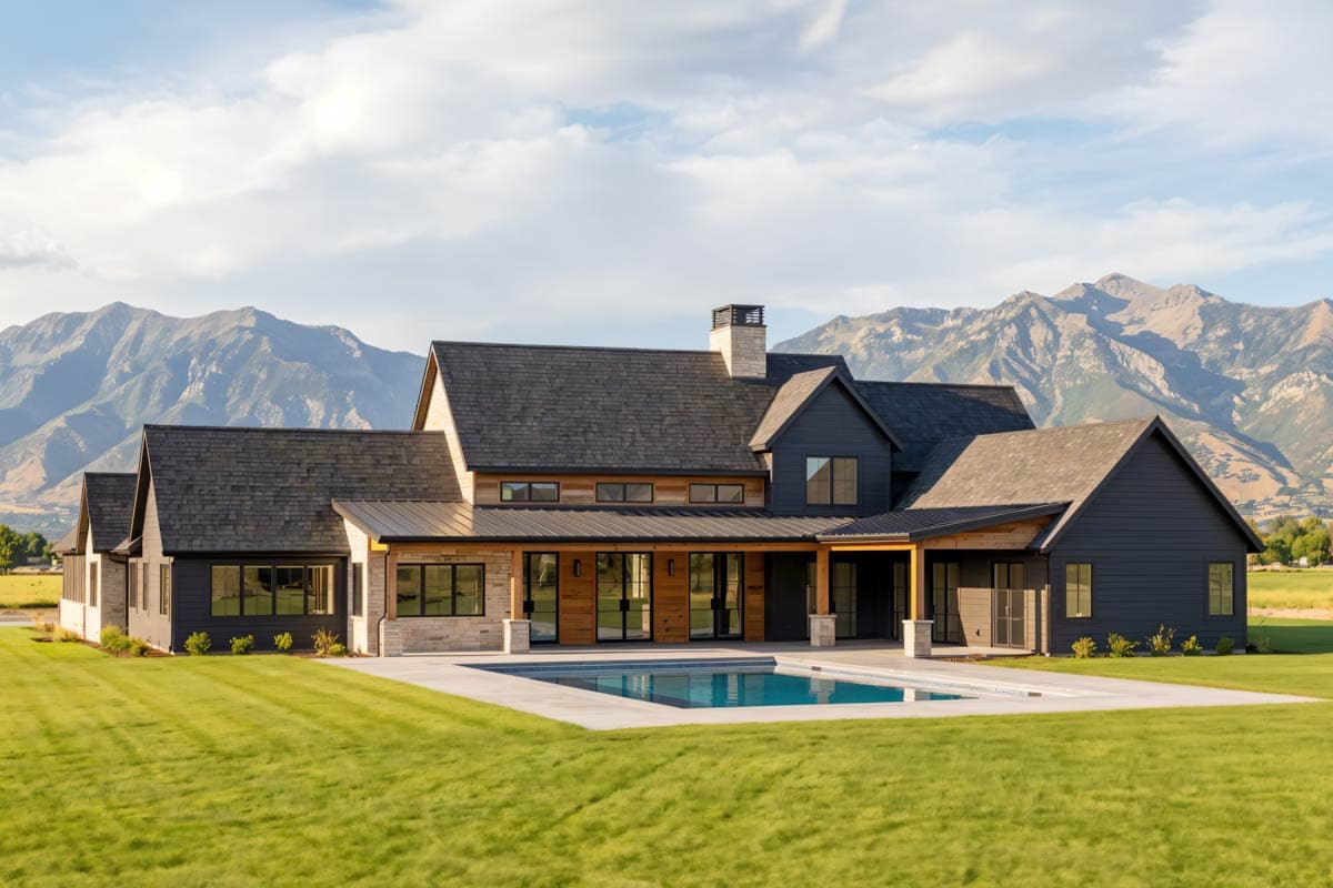 Modern farmhouse with a dark exterior, a swimming pool, and a backdrop of mountains under a partly cloudy sky.