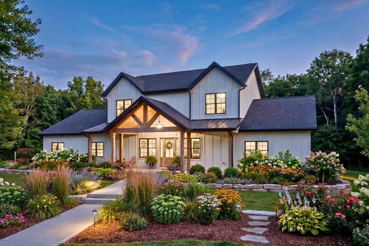 A two-story farmhouse with white siding and a dark roof, surrounded by lush landscaping and a pathway leading to the front door at dusk.