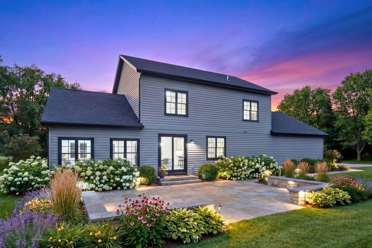 A two-story gray house with black trim, a stone patio, and vibrant landscaping under a dusk sky with pink and purple hues.