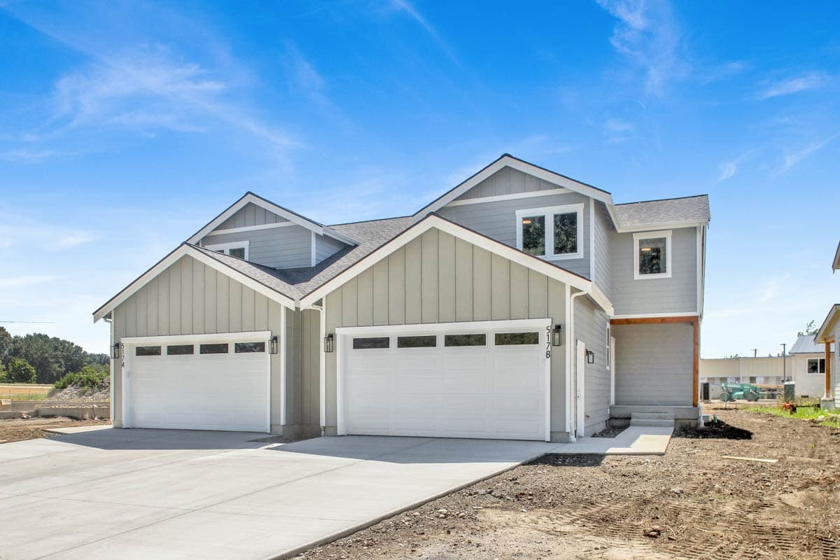 A newly constructed duplex with two white garage doors, grey siding, and a contrasting blue sky above. Construction site visible in the foreground.