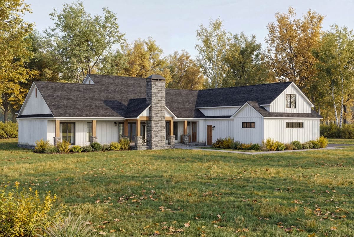 A white farmhouse with a dark roof and a stone chimney, set in a grassy yard surrounded by trees with autumn foliage.