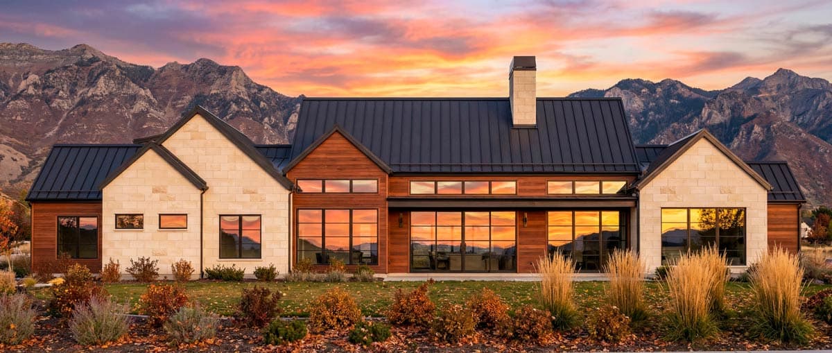 A modern home with wood and stone siding reflects a sunset sky with a mountain backdrop. Landscaping includes ornamental grass and fall foliage.