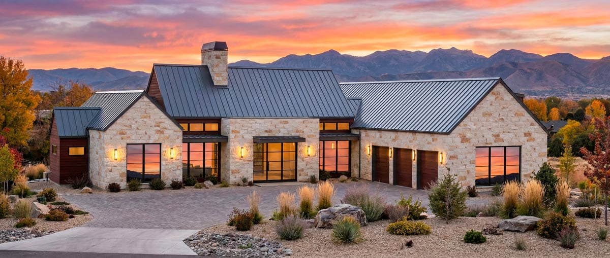 A modern stone house with a dark metal roof at sunset. Large windows reflect the colorful sky, and mountains are in the background.