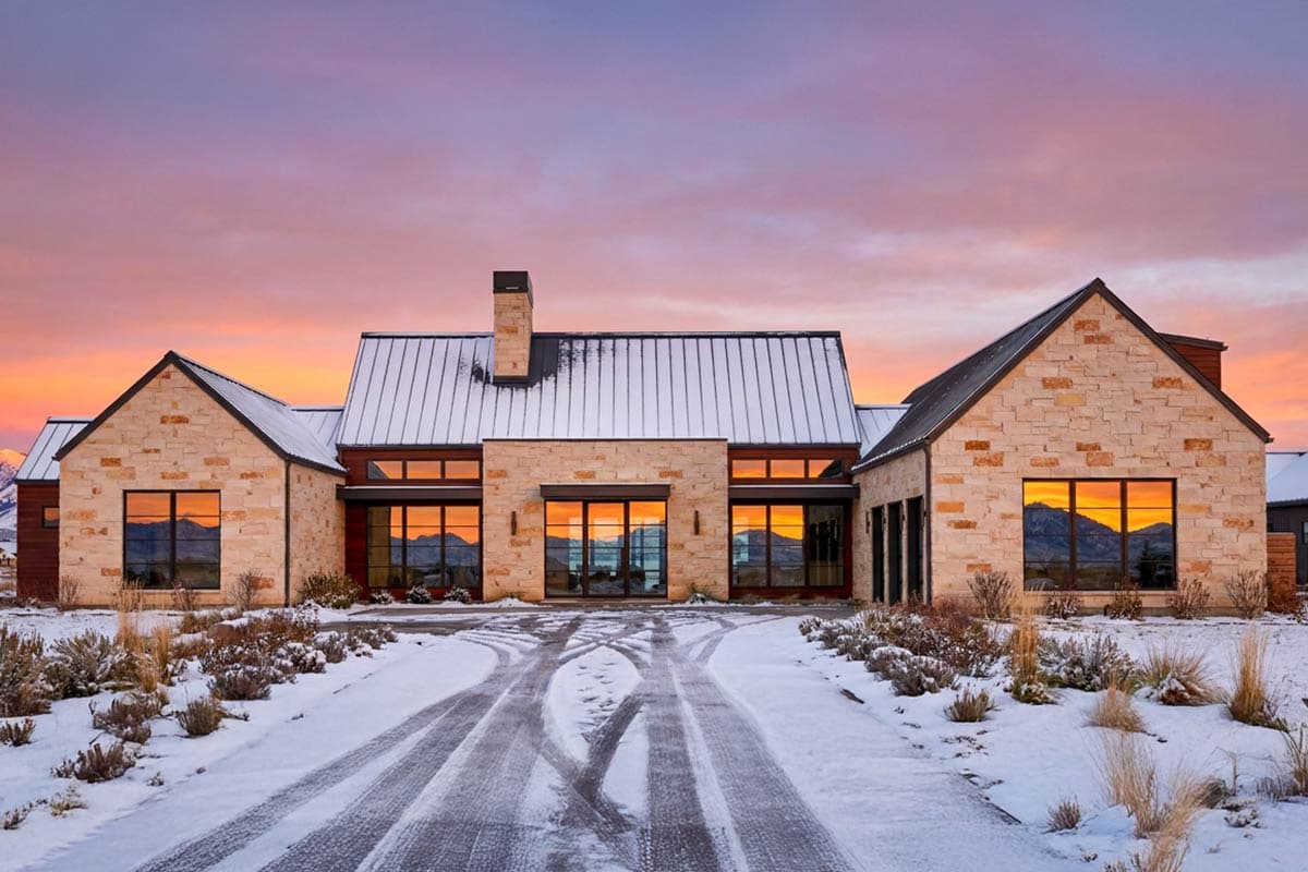 A modern stone house at sunset, with large windows reflecting the sky and mountains. Snow covers the roof and driveway. 