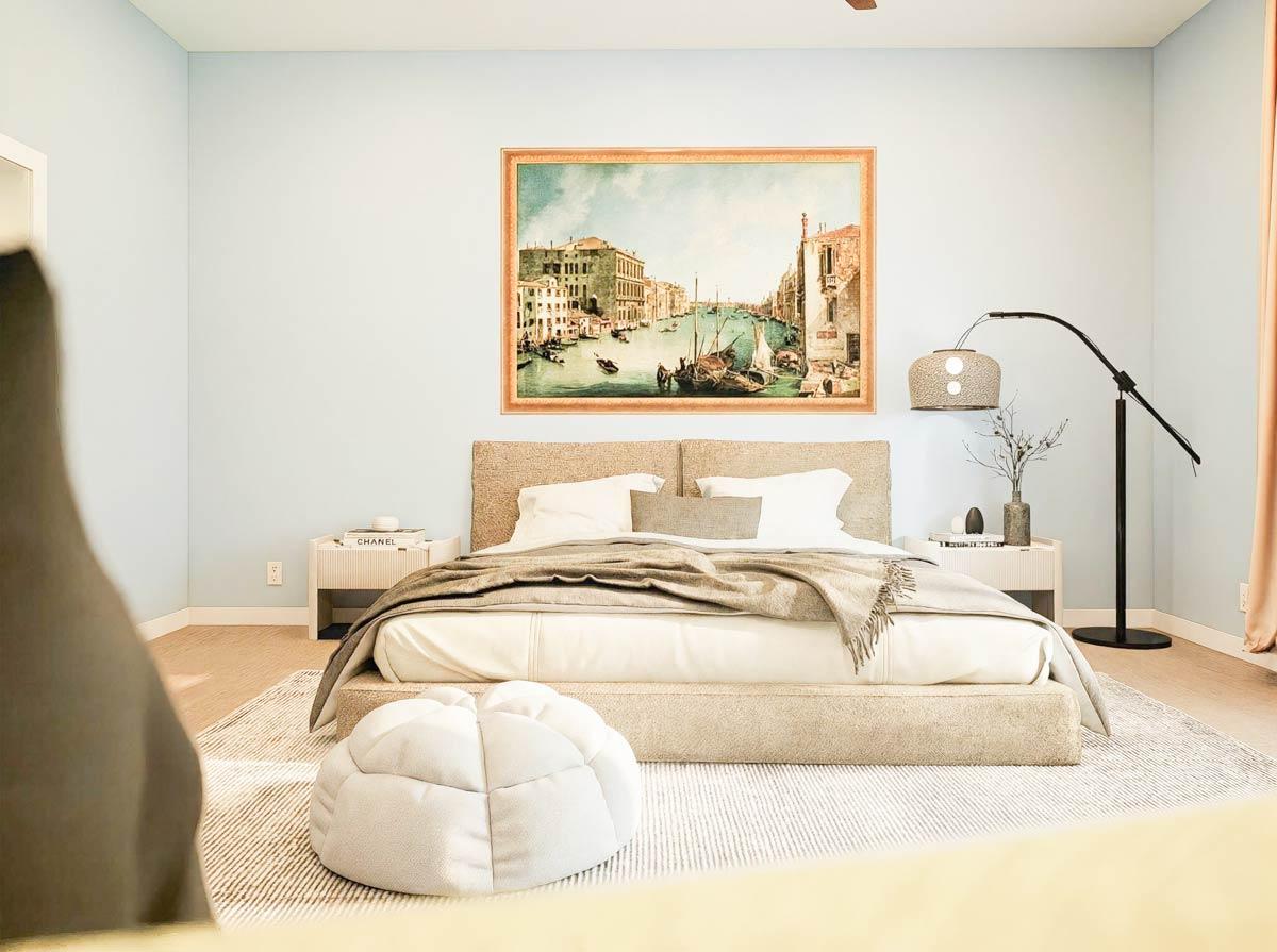 Bedroom with a framed canal print above a upholstered bed, bedside tables with lamps, and a floor pouf on a rug.
