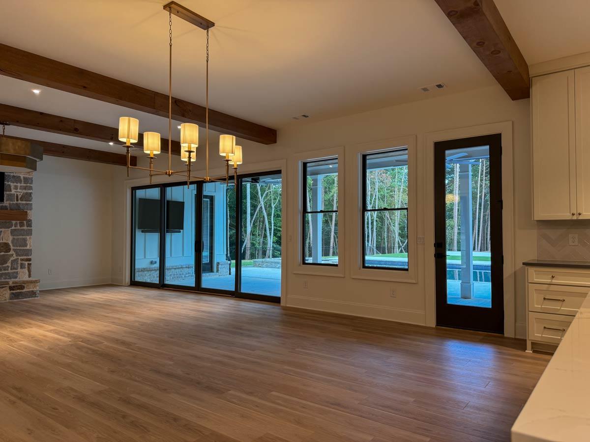 Open concept living space with wood beams, stone fireplace, large windows, and sliding glass doors leading to an outdoor pool area.