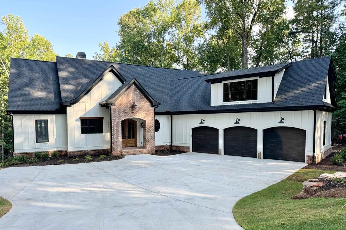A modern farmhouse with white siding, black trim, and a brick accent. A concrete driveway leads to the three-car garage and front door.
