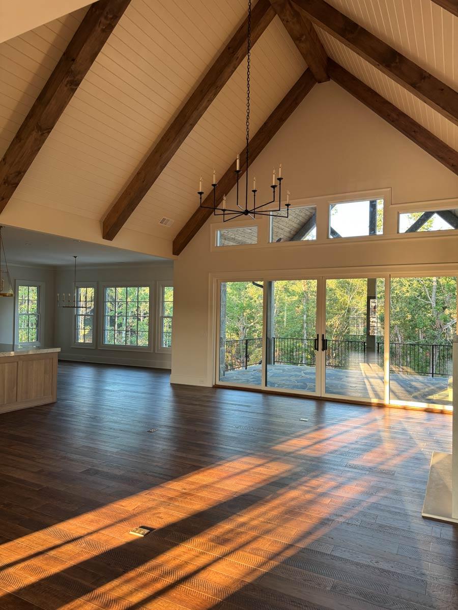 Interior view of open-concept living area with vaulted beamed ceiling, wood floors, and large windows leading to a deck.
