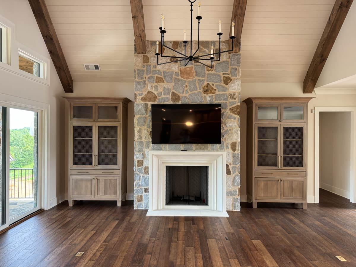Great room with vaulted wood beam ceiling, stone fireplace, built-in cabinets, and large windows.