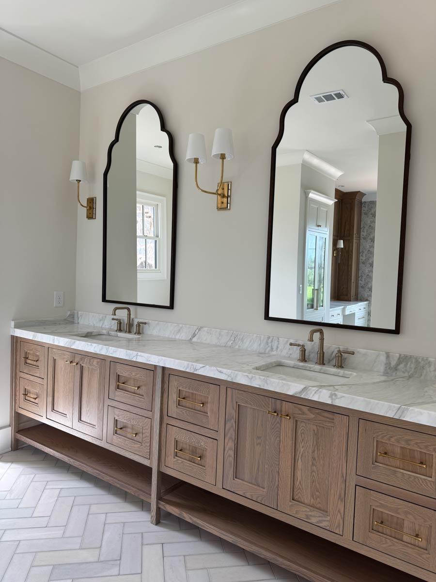 Bathroom vanity with marble countertop, double sinks, wood cabinets, and arched mirrors with sconces.