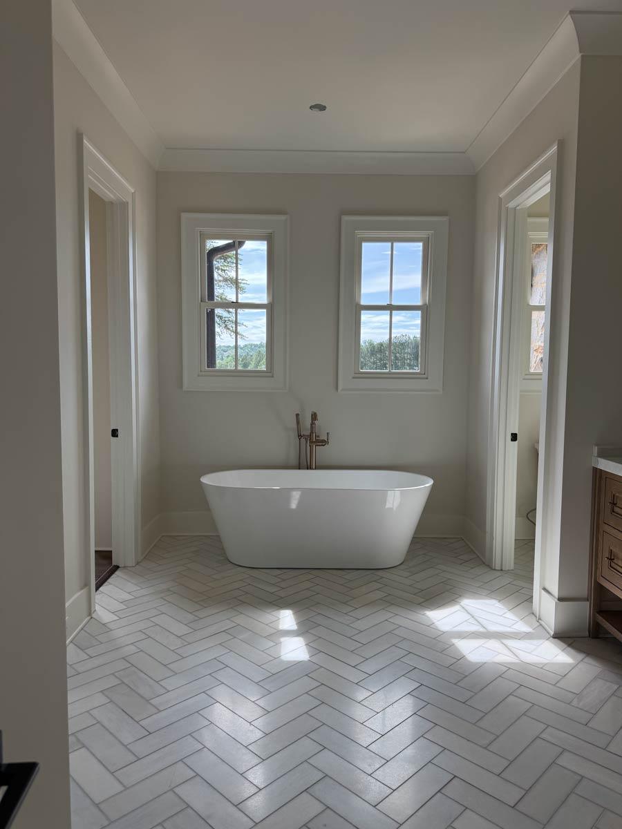Freestanding tub centered between two windows with herringbone tile floor and crown molding.